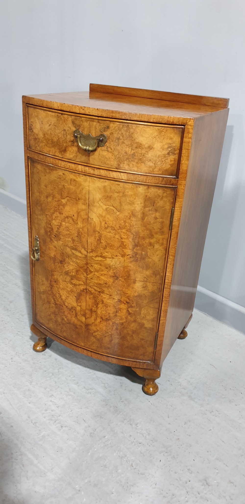 A compact bow-front burr walnut bedside cabinet featuring a single top drawer, lower cupboard, brass hardware, and short cabriole legs. View II