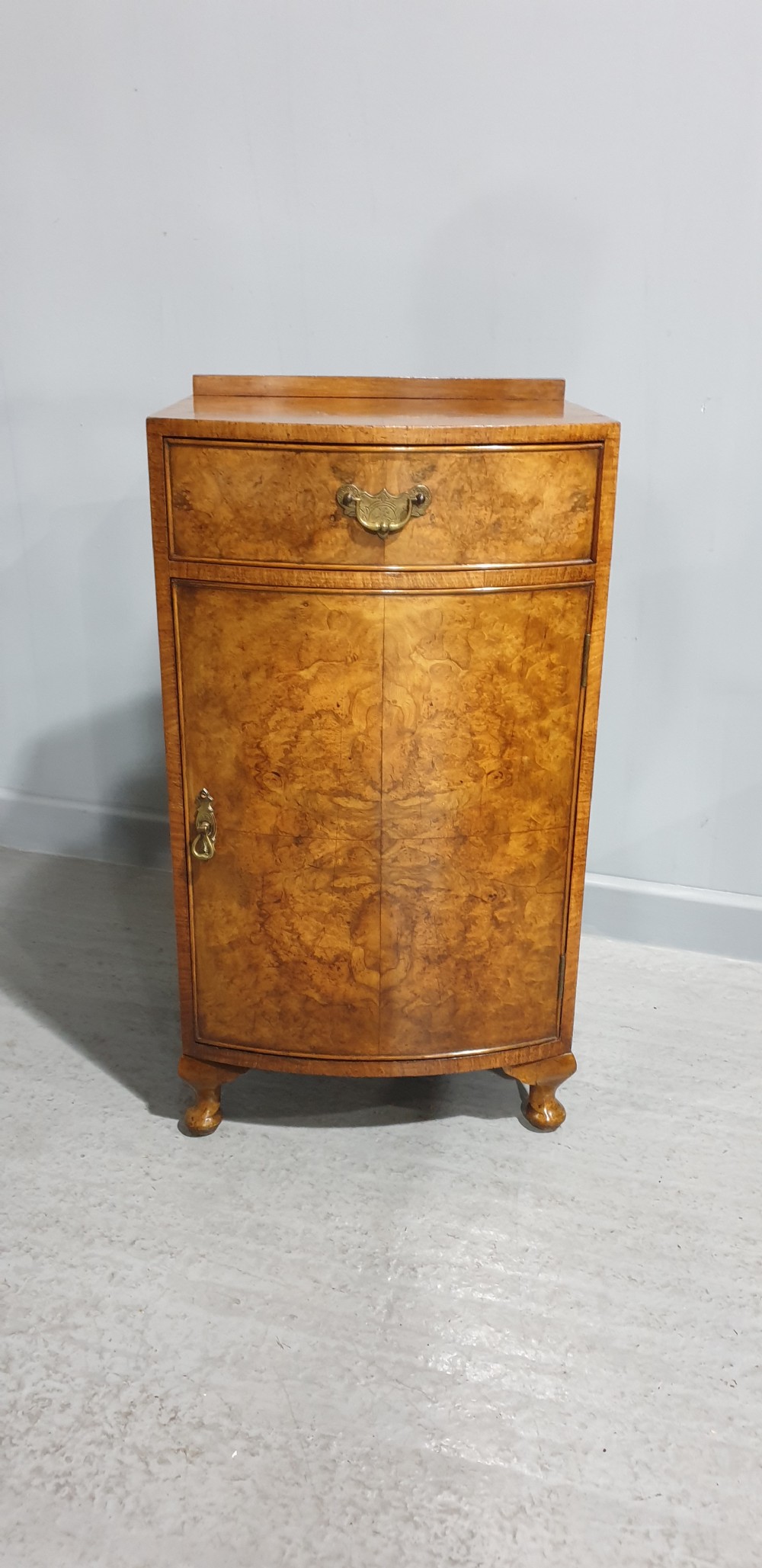 A compact bow-front burr walnut bedside cabinet featuring a single top drawer, lower cupboard, brass hardware, and short cabriole legs. View III