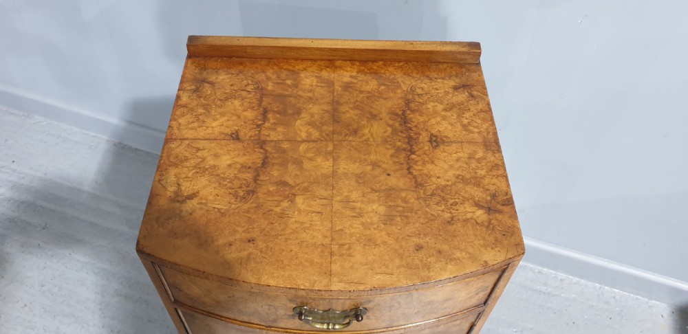 A compact bow-front burr walnut bedside cabinet featuring a single top drawer, lower cupboard, brass hardware, and short cabriole legs. View IV