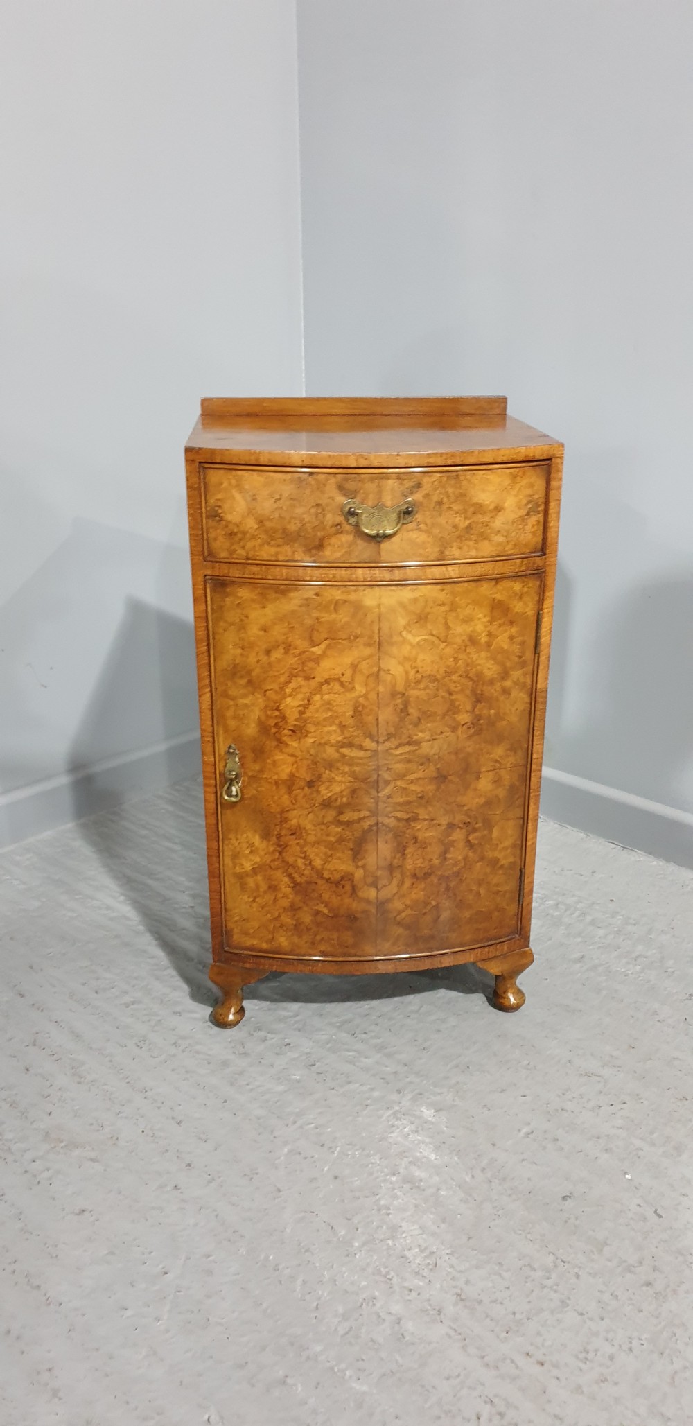 A compact bow-front burr walnut bedside cabinet featuring a single top drawer, lower cupboard, brass hardware, and short cabriole legs. View V