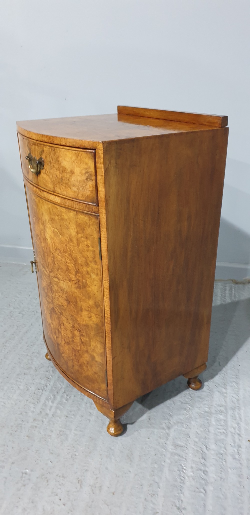 A compact bow-front burr walnut bedside cabinet featuring a single top drawer, lower cupboard, brass hardware, and short cabriole legs. View VII