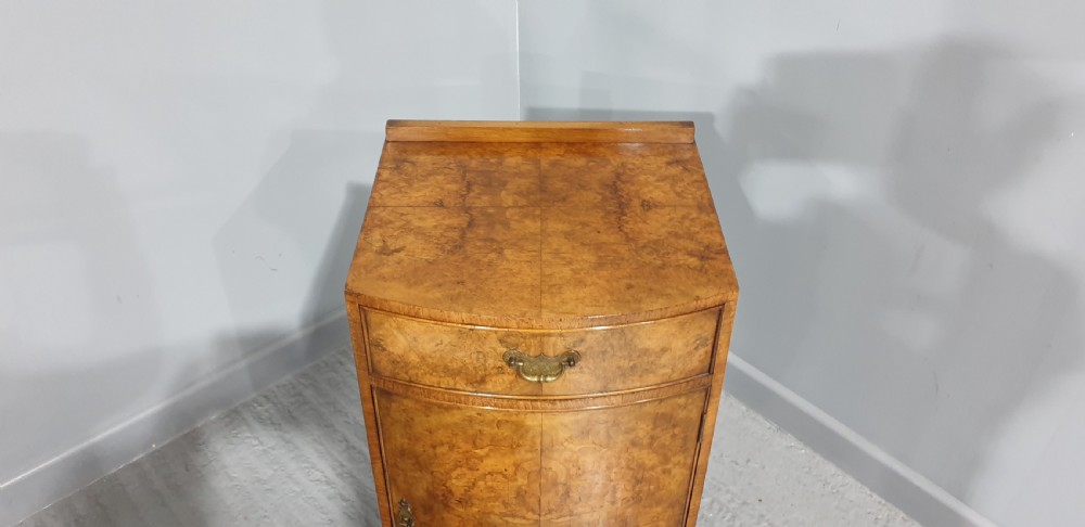 A compact bow-front burr walnut bedside cabinet featuring a single top drawer, lower cupboard, brass hardware, and short cabriole legs. View VIII