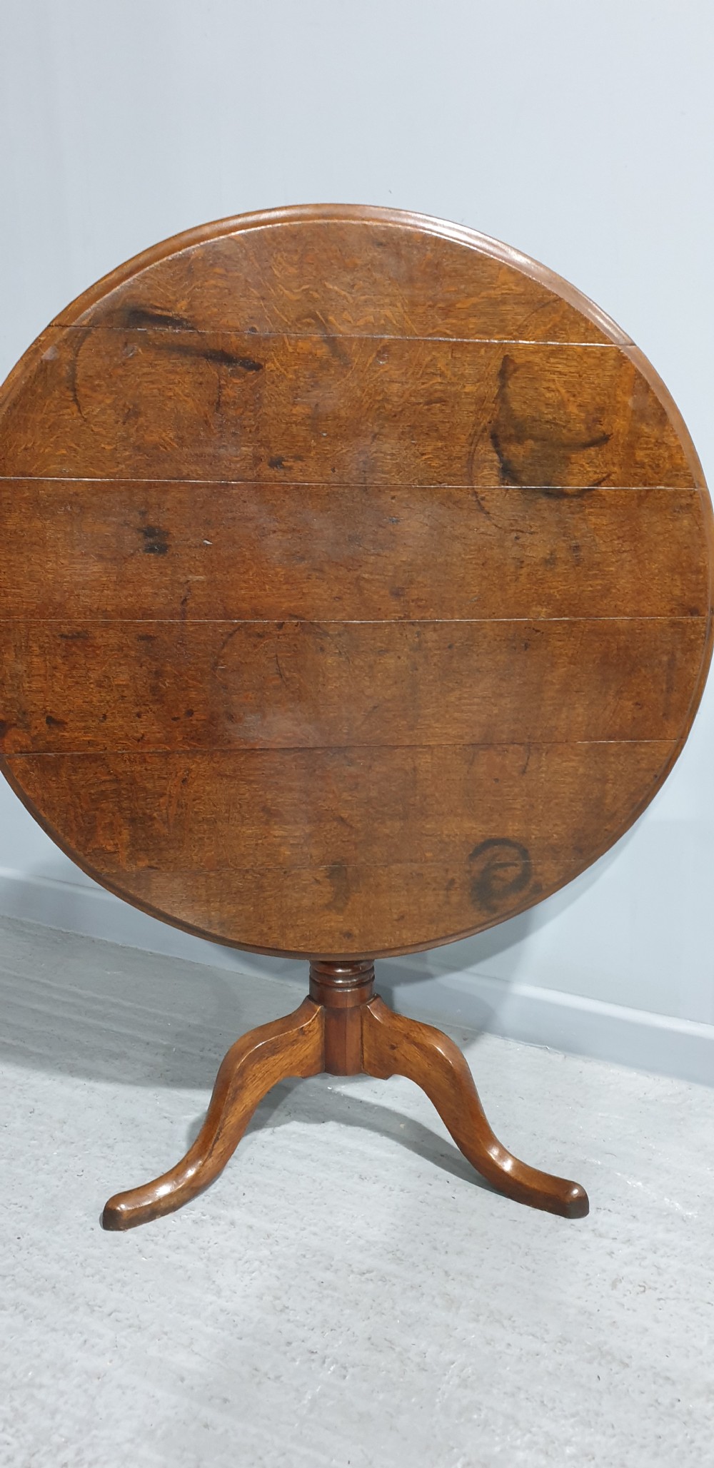 A Georgian period circular oak table shown with its tilt-top surface hinged vertically above a central turned pillar and three splayed cabriole legs. View II