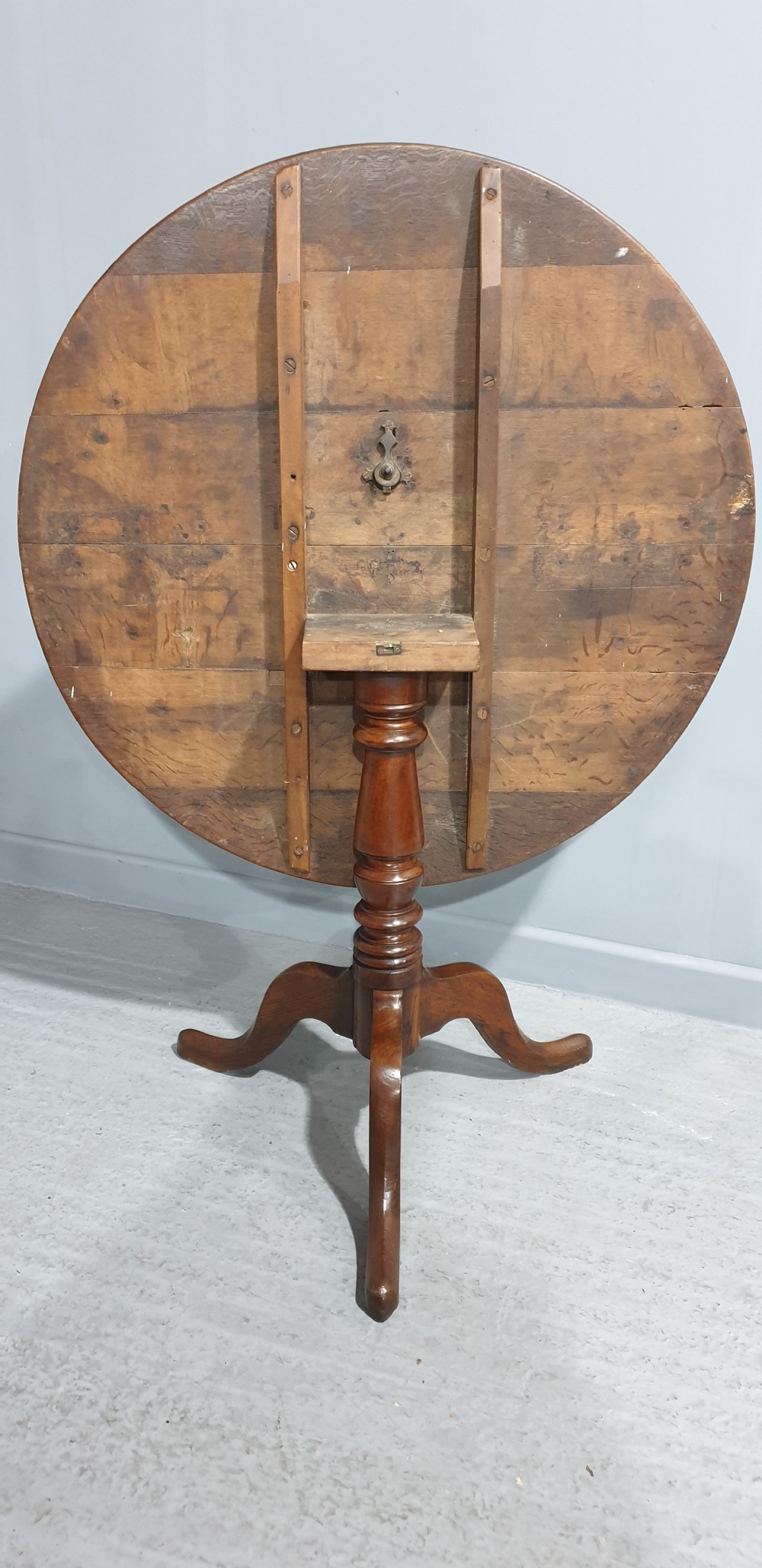 A Georgian period circular oak table shown with its tilt-top surface hinged vertically above a central turned pillar and three splayed cabriole legs. View III