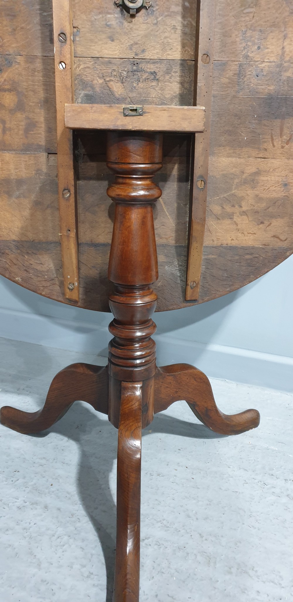 A Georgian period circular oak table shown with its tilt-top surface hinged vertically above a central turned pillar and three splayed cabriole legs. View IV