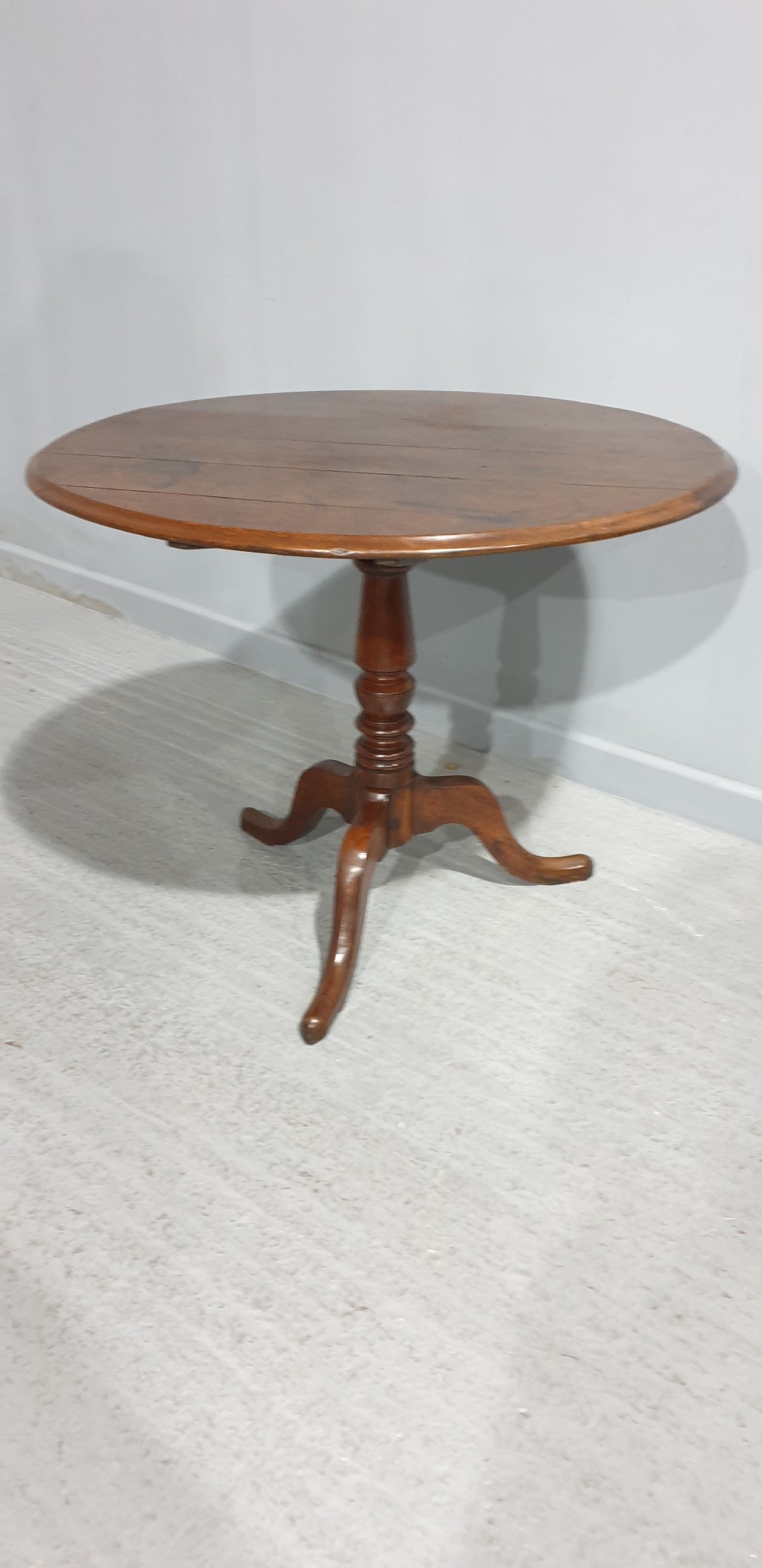 A Georgian period circular oak table shown with its tilt-top surface hinged vertically above a central turned pillar and three splayed cabriole legs. View V