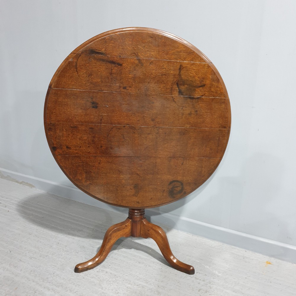 A Georgian period circular oak table shown with its tilt-top surface hinged vertically above a central turned pillar and three splayed cabriole legs.