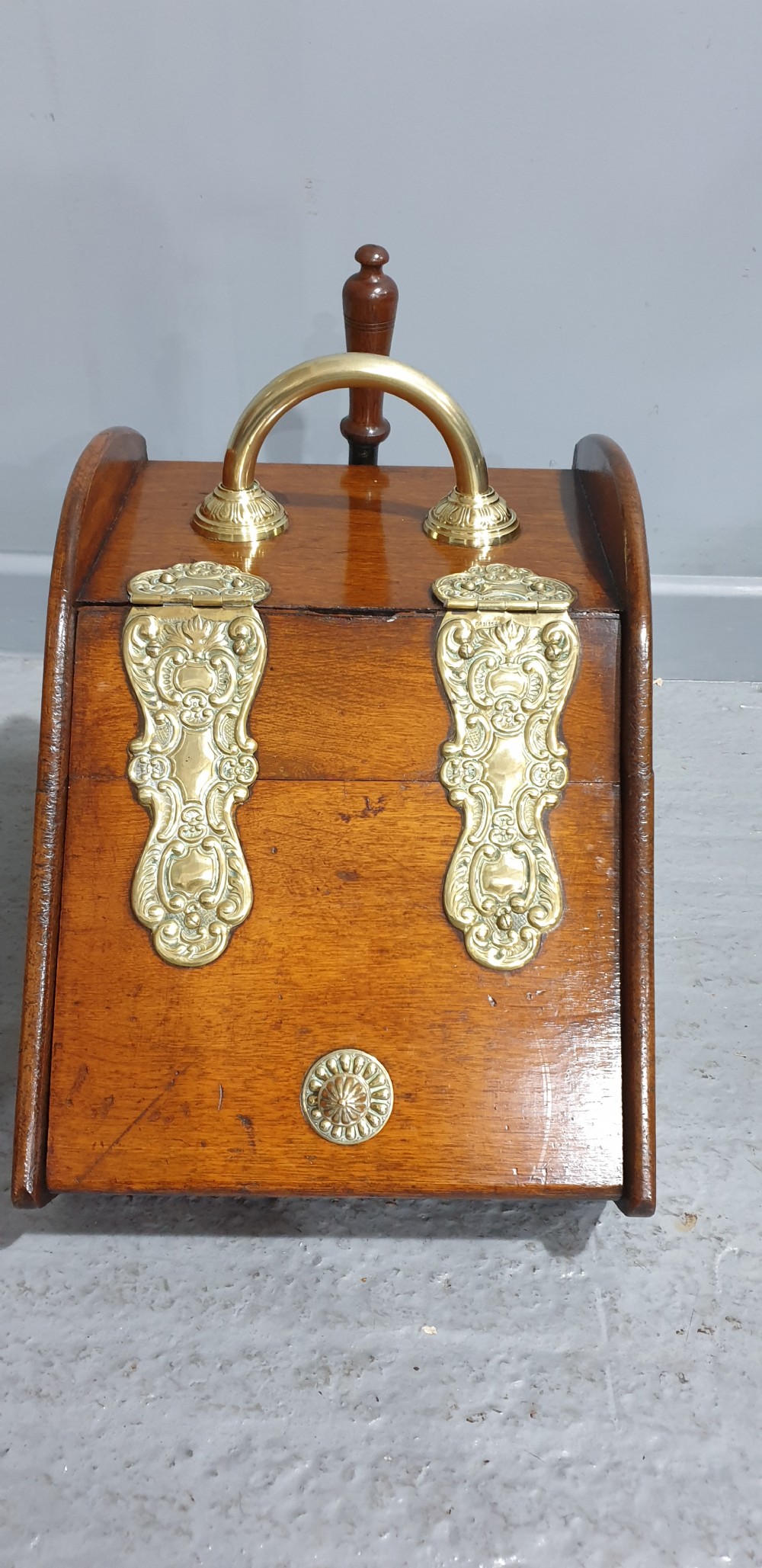 An antique oak coal scuttle featuring a sloped front flap with decorative brass hinges, a central brass carrying handle, and a wooden shovel handle at the rear. View II