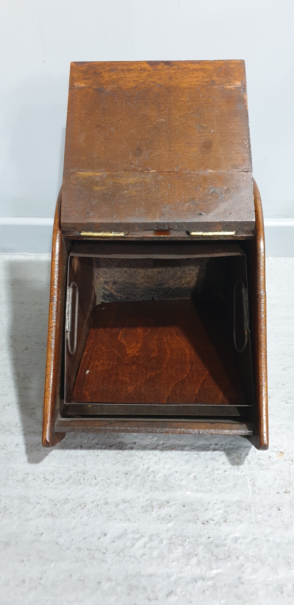 An antique oak coal scuttle featuring a sloped front flap with decorative brass hinges, a central brass carrying handle, and a wooden shovel handle at the rear. View III
