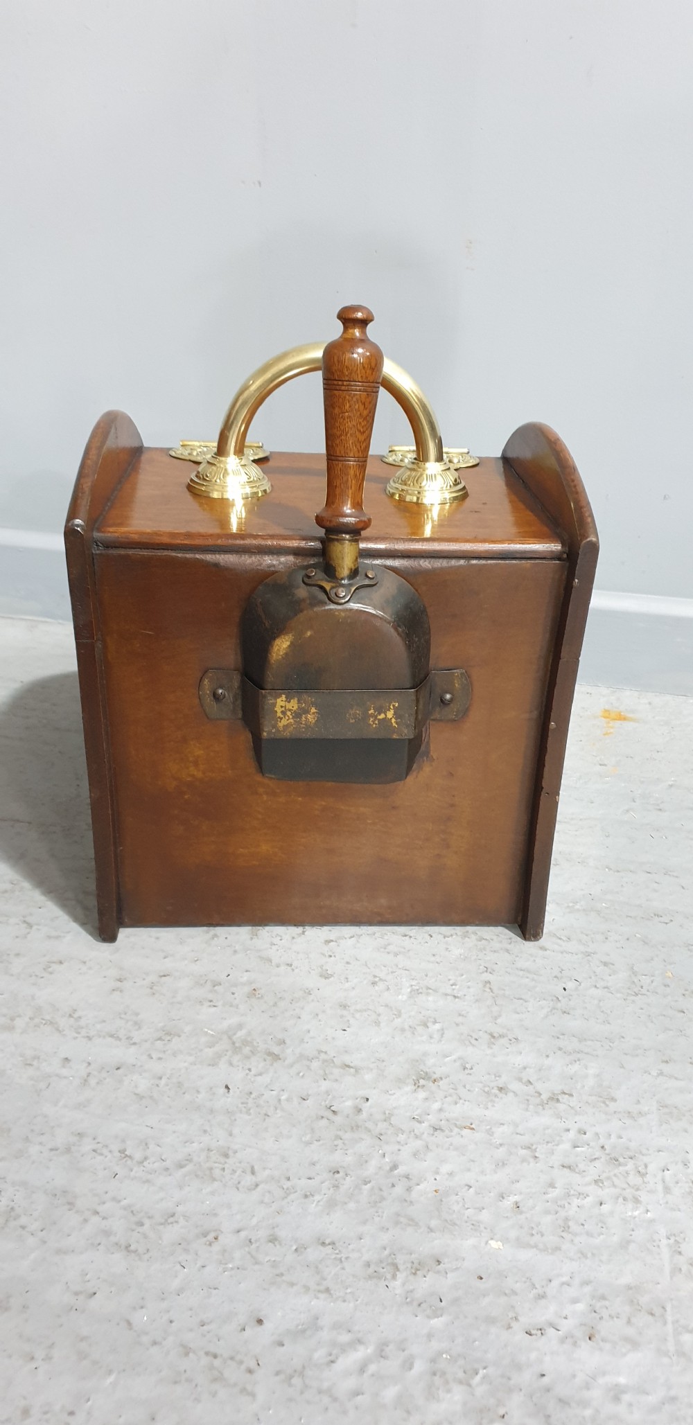 An antique oak coal scuttle featuring a sloped front flap with decorative brass hinges, a central brass carrying handle, and a wooden shovel handle at the rear. View IV