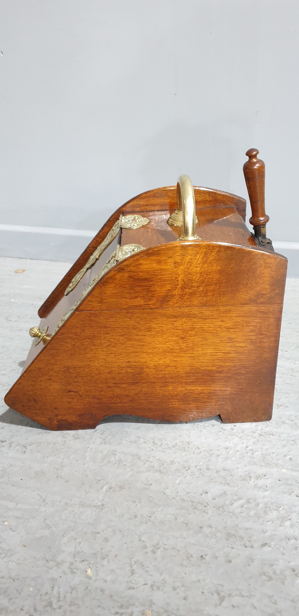 An antique oak coal scuttle featuring a sloped front flap with decorative brass hinges, a central brass carrying handle, and a wooden shovel handle at the rear. View V