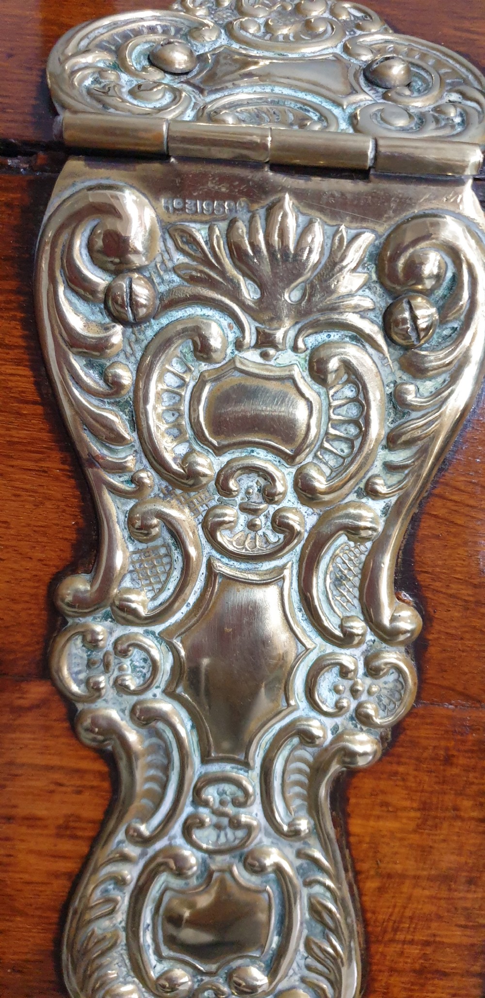 An antique oak coal scuttle featuring a sloped front flap with decorative brass hinges, a central brass carrying handle, and a wooden shovel handle at the rear. View VI