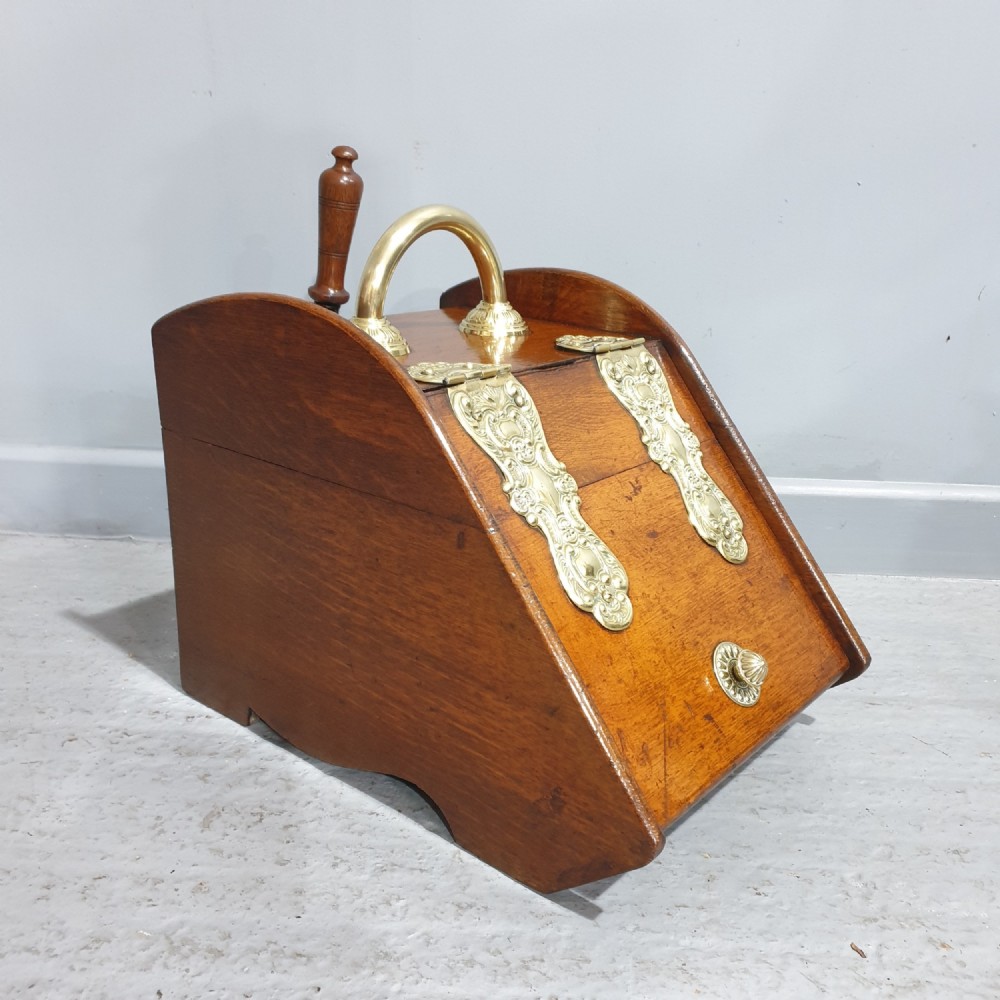 An antique oak coal scuttle featuring a sloped front flap with decorative brass hinges, a central brass carrying handle, and a wooden shovel handle at the rear.