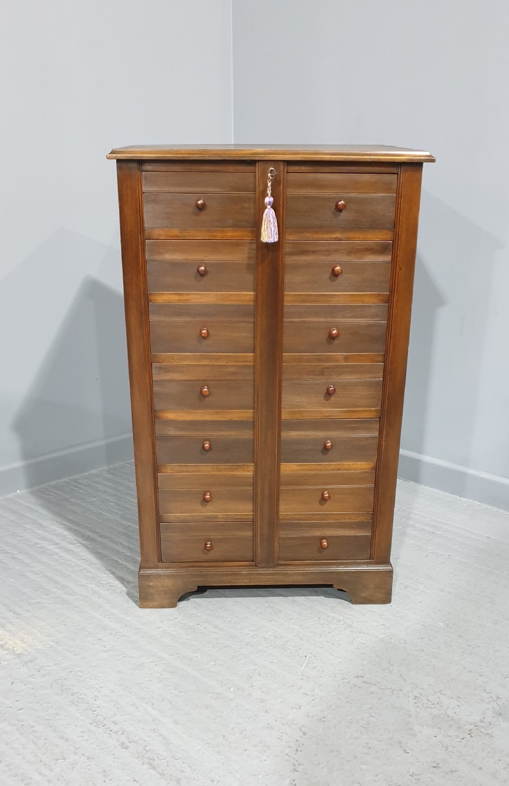 A tall warm brown wooden specimen chest of drawers featuring seven shallow drawers with turned knobs and a central vertical locking bar.