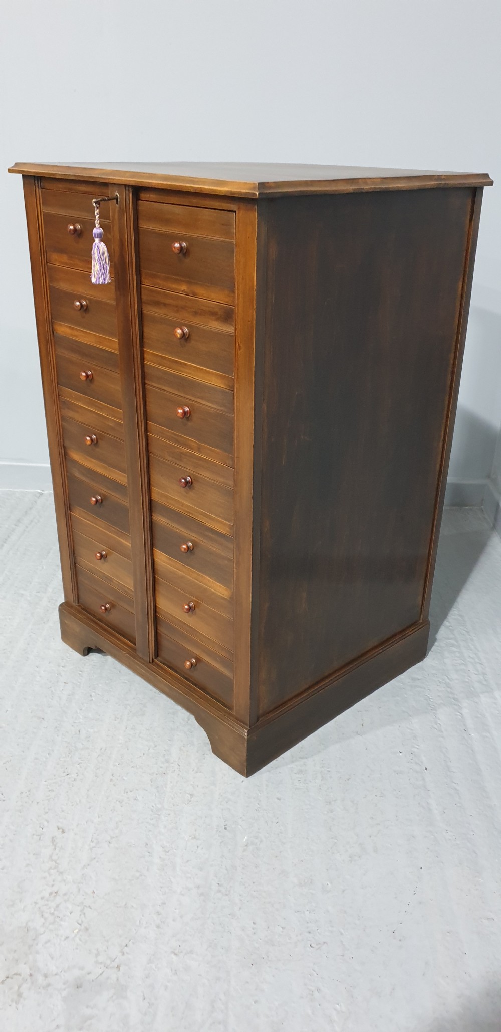 A tall warm brown wooden specimen chest of drawers featuring seven shallow drawers with turned knobs and a central vertical locking bar. View III