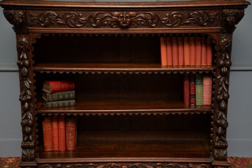 fig 2. A low solid oak open bookcase decorated with hand-carved Green Man motifs, foliate pilasters, three shelves with scalloped edges, and a plinth base.