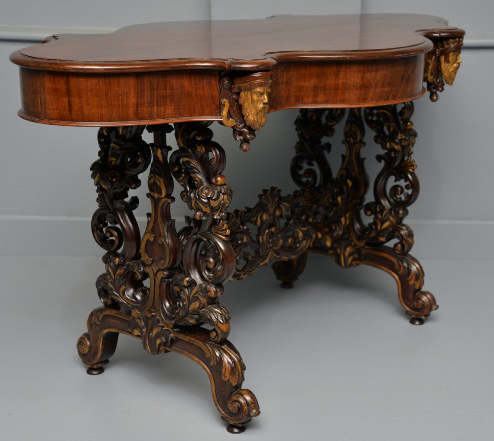 A 19th-century dark carved mahogany centre table featuring a shaped top, parcel-gilt mask corner details, and intricate openwork scroll legs.