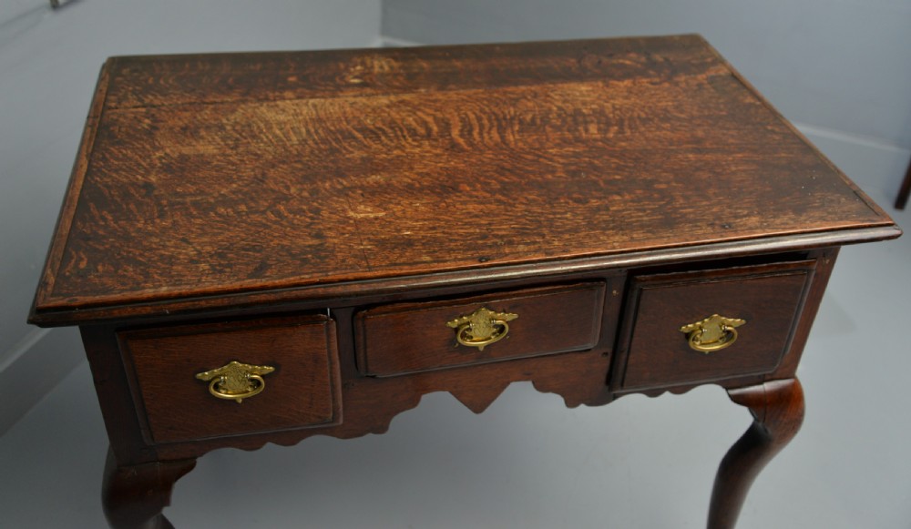 An early eighteenth-century oak lowboy featuring three drawers with brass handles, a scalloped apron, and four cabriole legs with pad feet. View XX