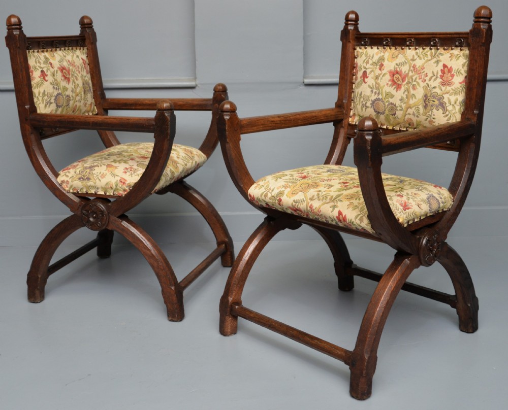 A pair of Victorian Gothic Revival dark oak X-frame armchairs featuring carved rosettes and floral tapestry upholstered seats and backrests with brass tack trimming.