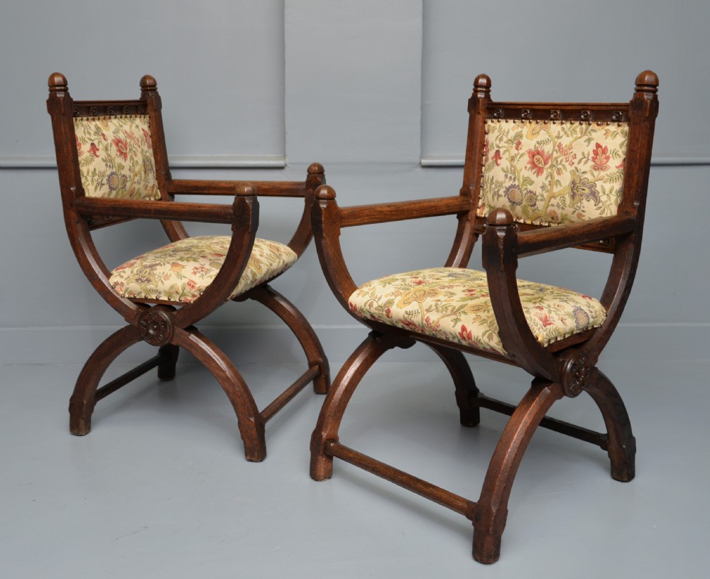 A pair of Victorian Gothic Revival dark oak X-frame armchairs featuring carved rosettes and floral tapestry upholstered seats and backrests with brass tack trimming. View II