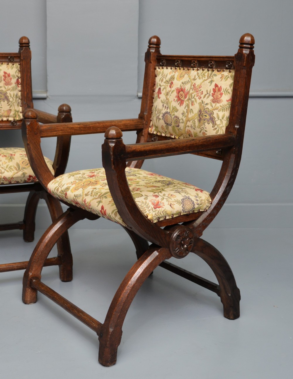 A pair of Victorian Gothic Revival dark oak X-frame armchairs featuring carved rosettes and floral tapestry upholstered seats and backrests with brass tack trimming. View V