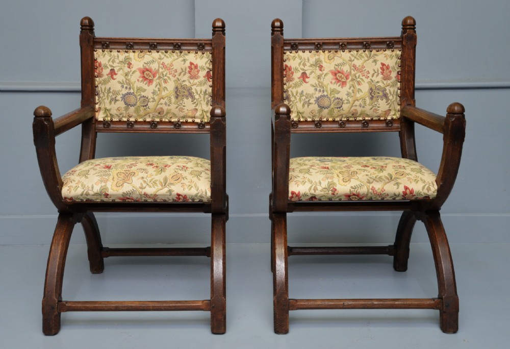 A pair of Victorian Gothic Revival dark oak X-frame armchairs featuring carved rosettes and floral tapestry upholstered seats and backrests with brass tack trimming. View VI
