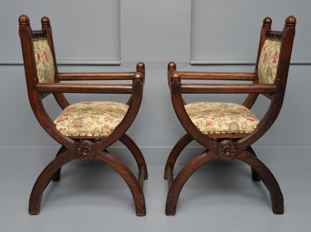 A pair of Victorian Gothic Revival dark oak X-frame armchairs featuring carved rosettes and floral tapestry upholstered seats and backrests with brass tack trimming. View VII