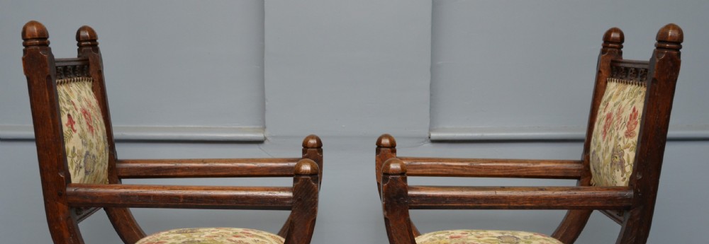 A pair of Victorian Gothic Revival dark oak X-frame armchairs featuring carved rosettes and floral tapestry upholstered seats and backrests with brass tack trimming. View IX