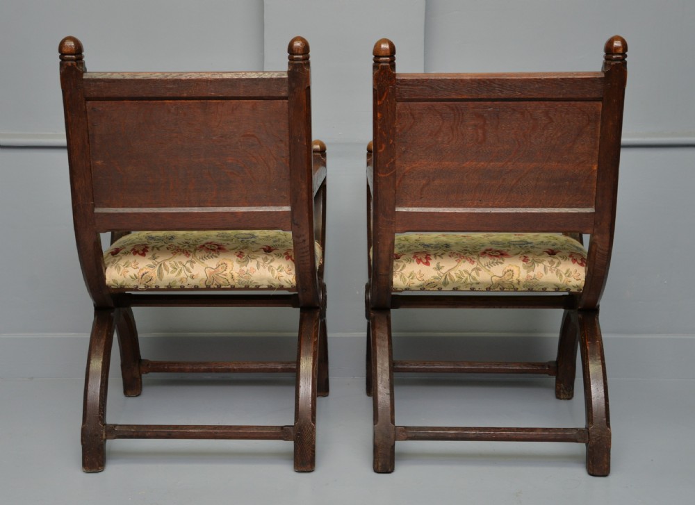A pair of Victorian Gothic Revival dark oak X-frame armchairs featuring carved rosettes and floral tapestry upholstered seats and backrests with brass tack trimming. View X