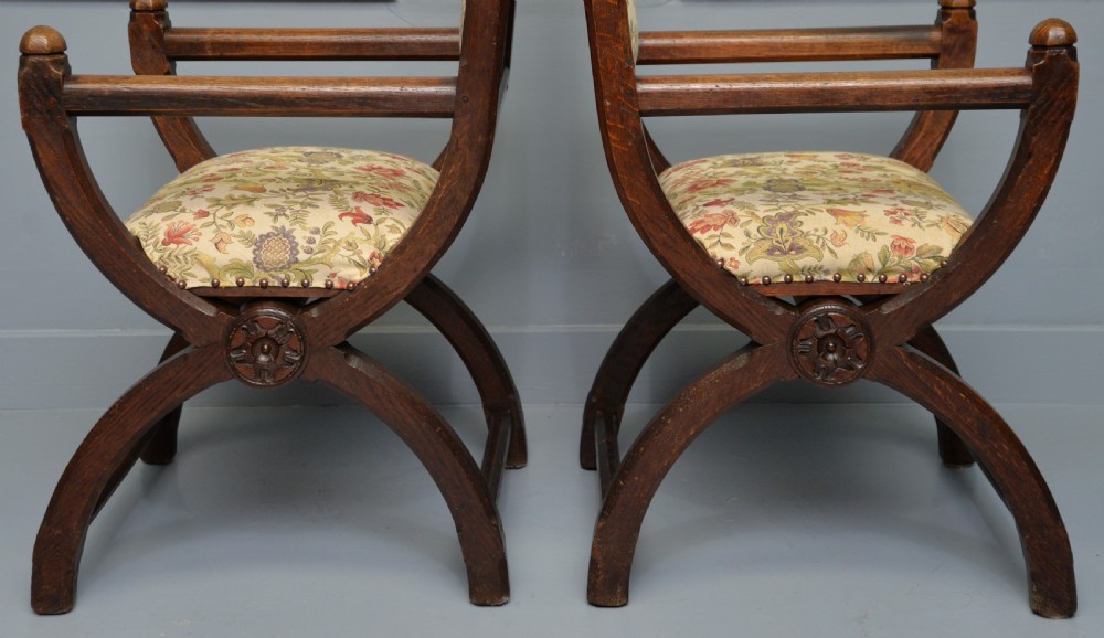 A pair of Victorian Gothic Revival dark oak X-frame armchairs featuring carved rosettes and floral tapestry upholstered seats and backrests with brass tack trimming. View XII