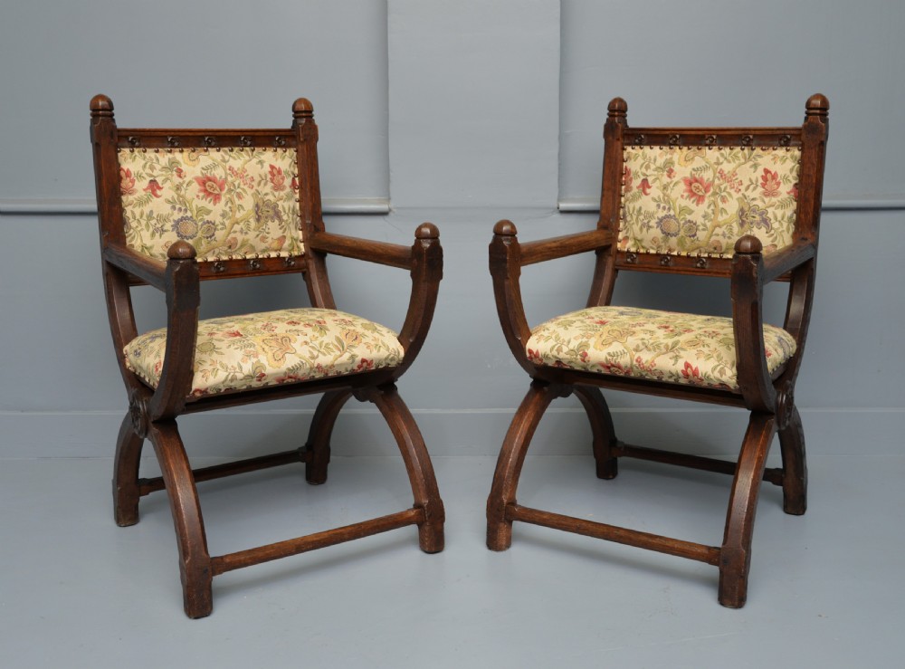 A pair of Victorian Gothic Revival dark oak X-frame armchairs featuring carved rosettes and floral tapestry upholstered seats and backrests with brass tack trimming. View XIII