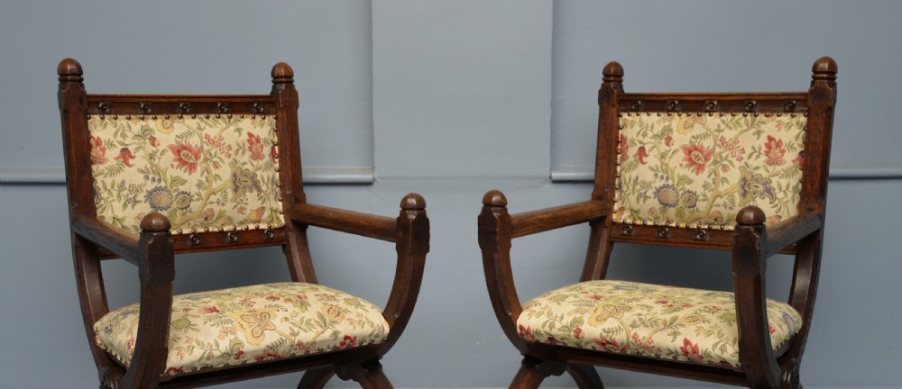 A pair of Victorian Gothic Revival dark oak X-frame armchairs featuring carved rosettes and floral tapestry upholstered seats and backrests with brass tack trimming. View XIV