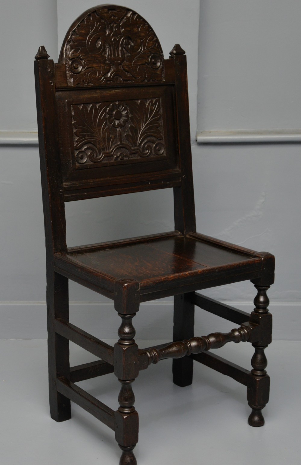 A dark carved oak 17th-century style backstool chair featuring an arched carved cresting rail, a carved floral back panel, turned front legs, and stretchers.