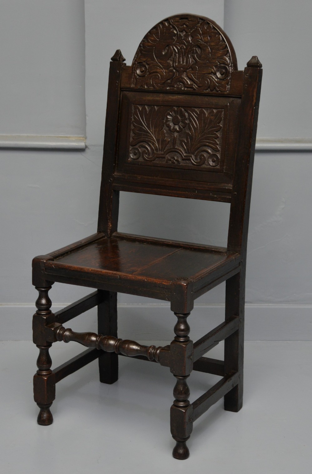 A dark carved oak 17th-century style backstool chair featuring an arched carved cresting rail, a carved floral back panel, turned front legs, and stretchers. View XVI
