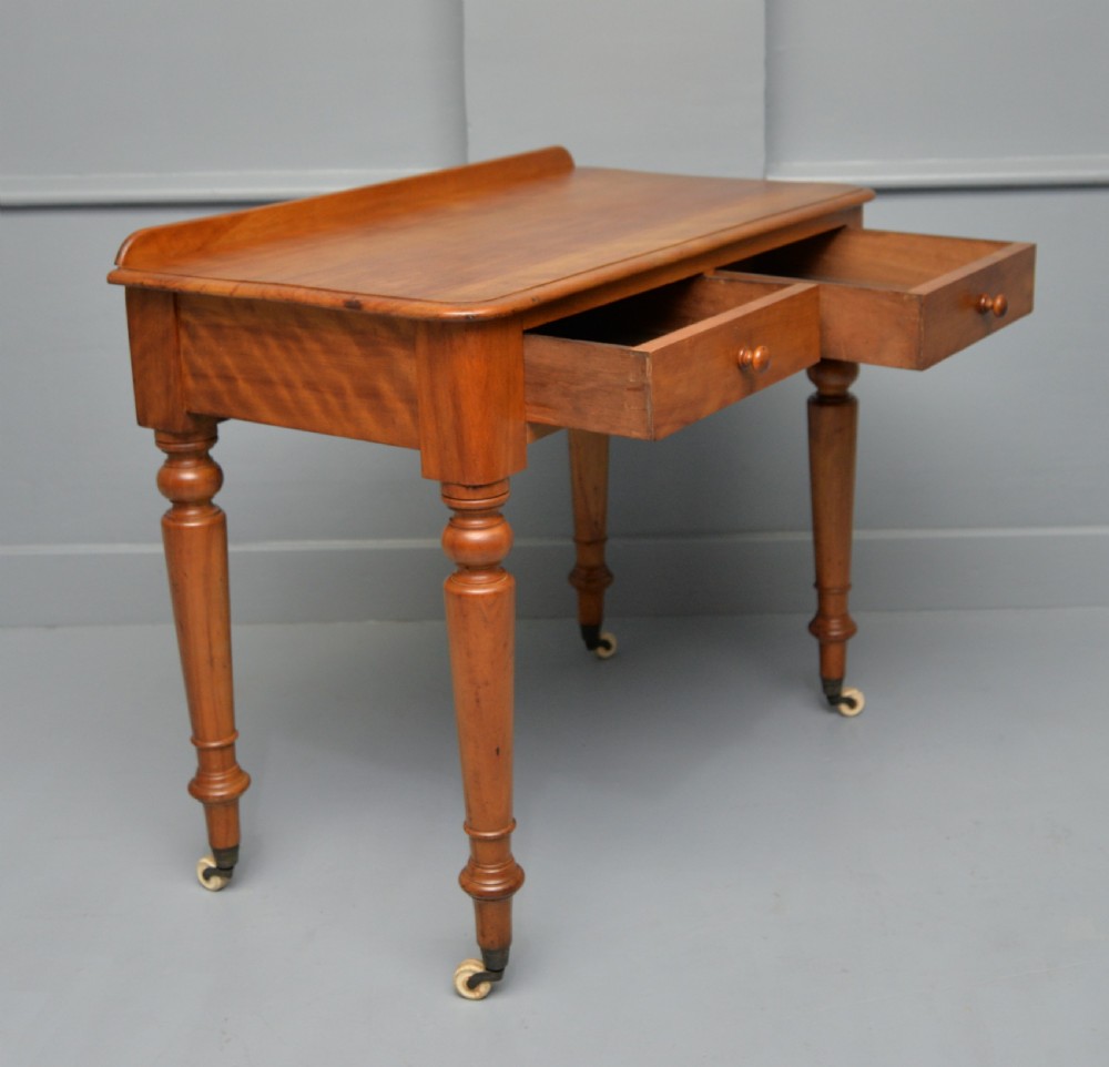 A Victorian satinwood writing table featuring two frieze drawers with wooden knobs, a low rear gallery, turned legs, and ceramic castors. View VI