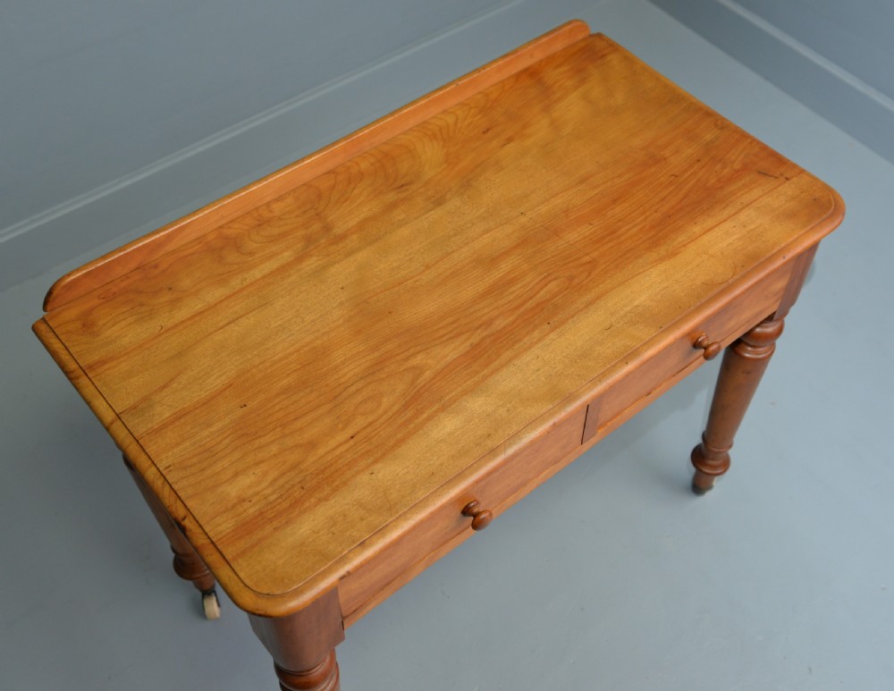 A Victorian satinwood writing table featuring two frieze drawers with wooden knobs, a low rear gallery, turned legs, and ceramic castors. View X