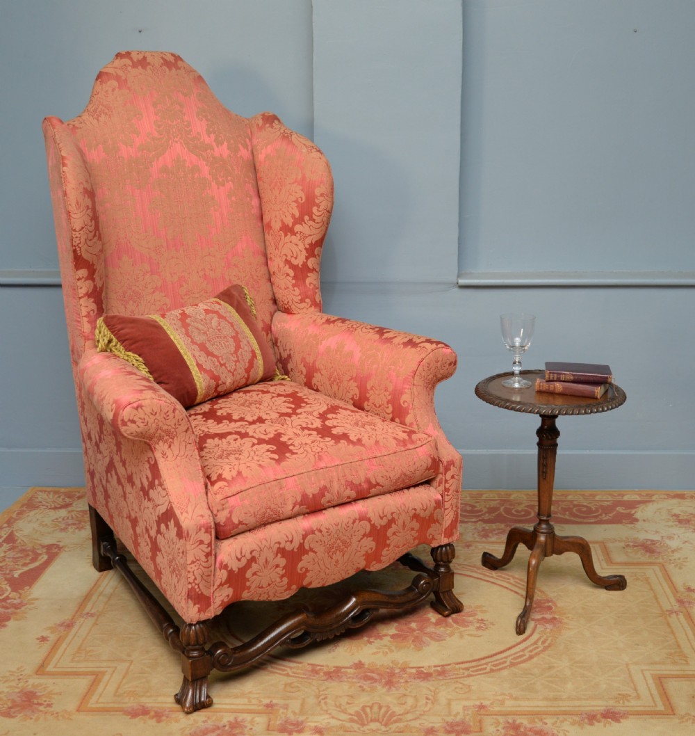 A high-backed wing armchair upholstered in patterned terracotta damask with a matching lumbar cushion and carved dark wood legs.