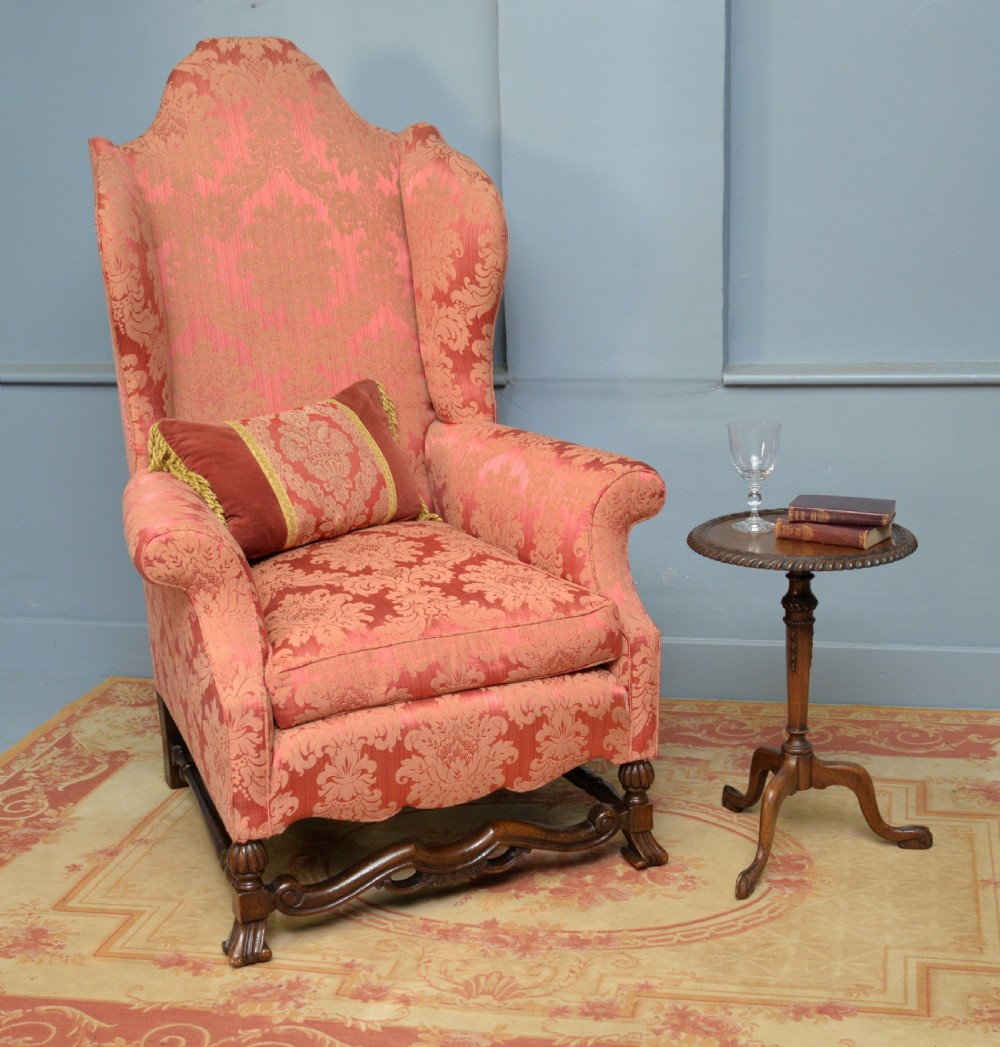 A high-backed wing armchair upholstered in patterned terracotta damask with a matching lumbar cushion and carved dark wood legs. View VI