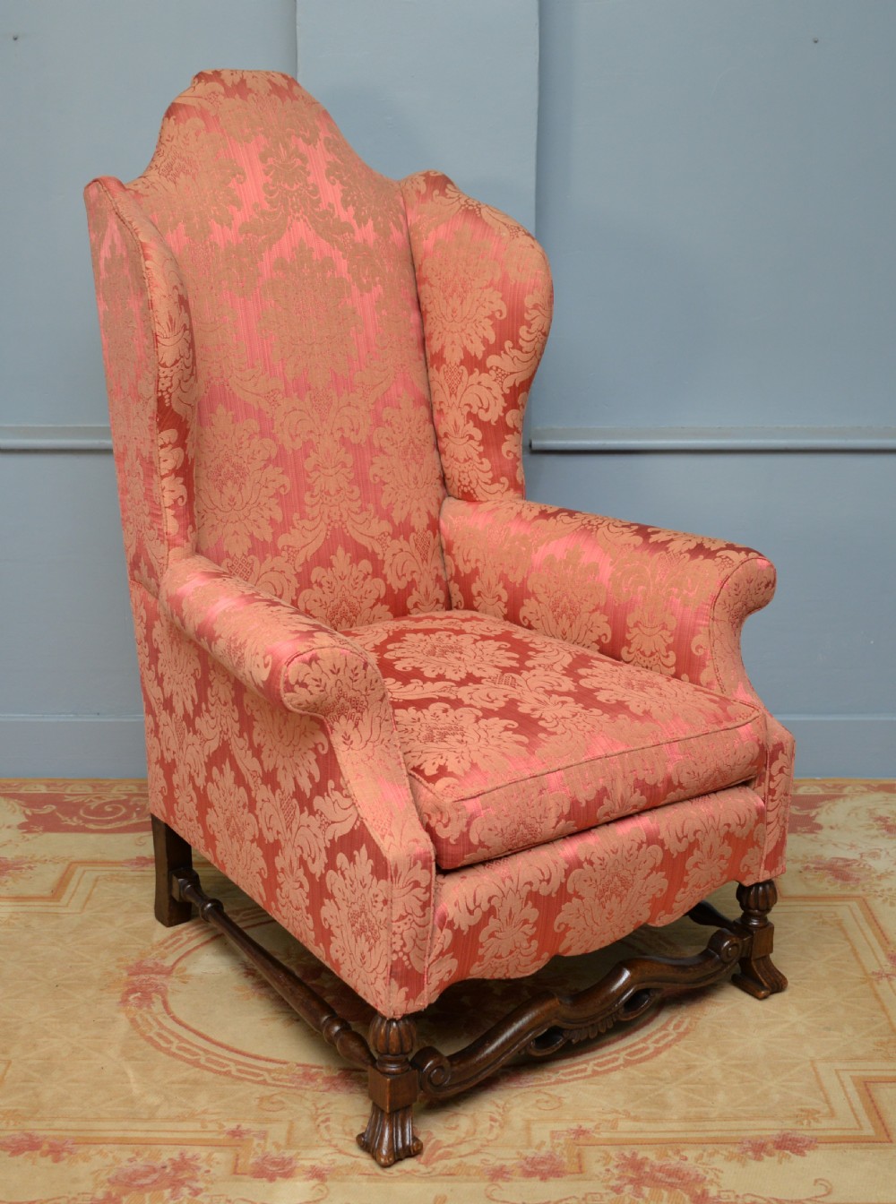 A high-backed wing armchair upholstered in patterned terracotta damask with a matching lumbar cushion and carved dark wood legs. View X