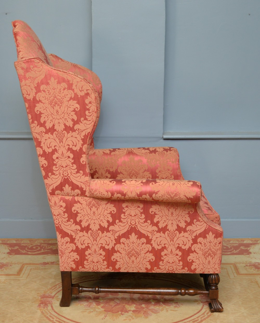 A high-backed wing armchair upholstered in patterned terracotta damask with a matching lumbar cushion and carved dark wood legs. View XII