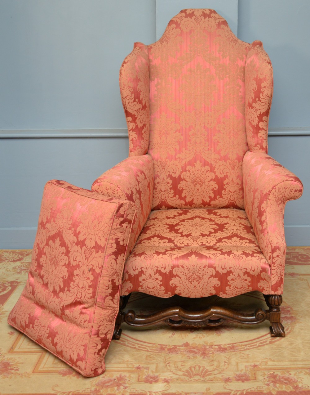 A high-backed wing armchair upholstered in patterned terracotta damask with a matching lumbar cushion and carved dark wood legs. View XIX