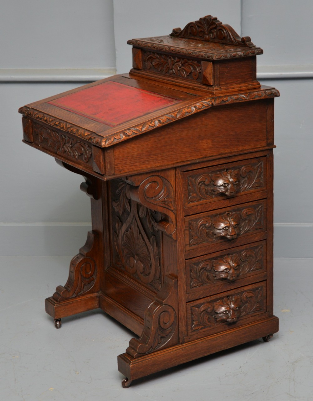 A Victorian dark carved oak Davenport desk featuring a red leather writing slant, carved Green Man mask handles on four side drawers, and a raised stationery gallery.