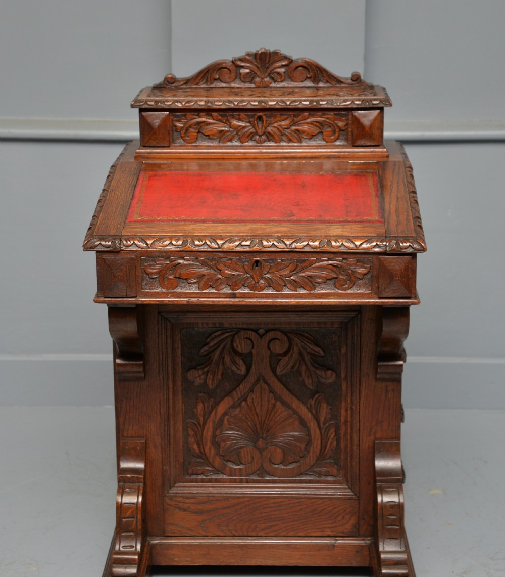A Victorian dark carved oak Davenport desk featuring a red leather writing slant, carved Green Man mask handles on four side drawers, and a raised stationery gallery. View III