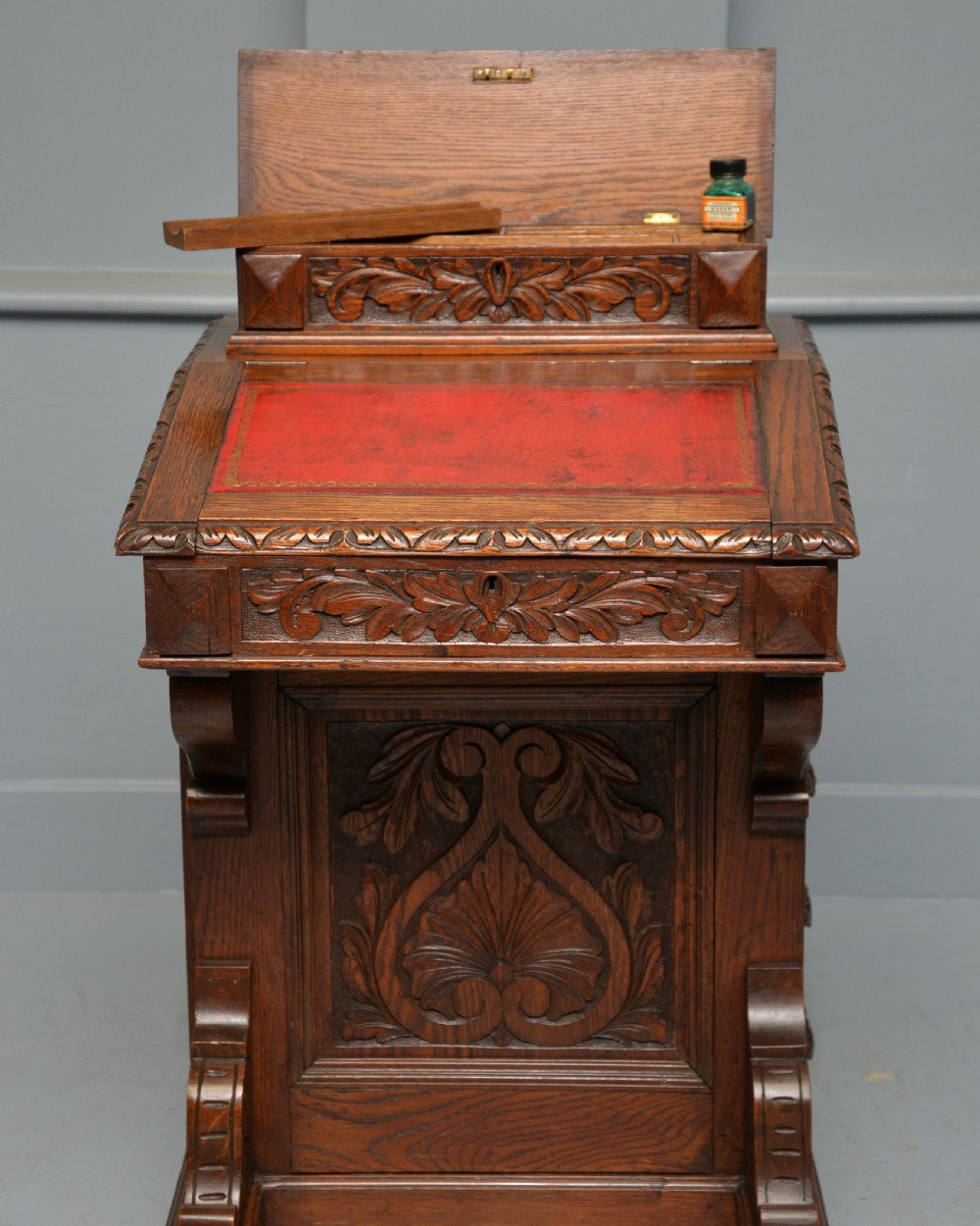 A Victorian dark carved oak Davenport desk featuring a red leather writing slant, carved Green Man mask handles on four side drawers, and a raised stationery gallery. View VI