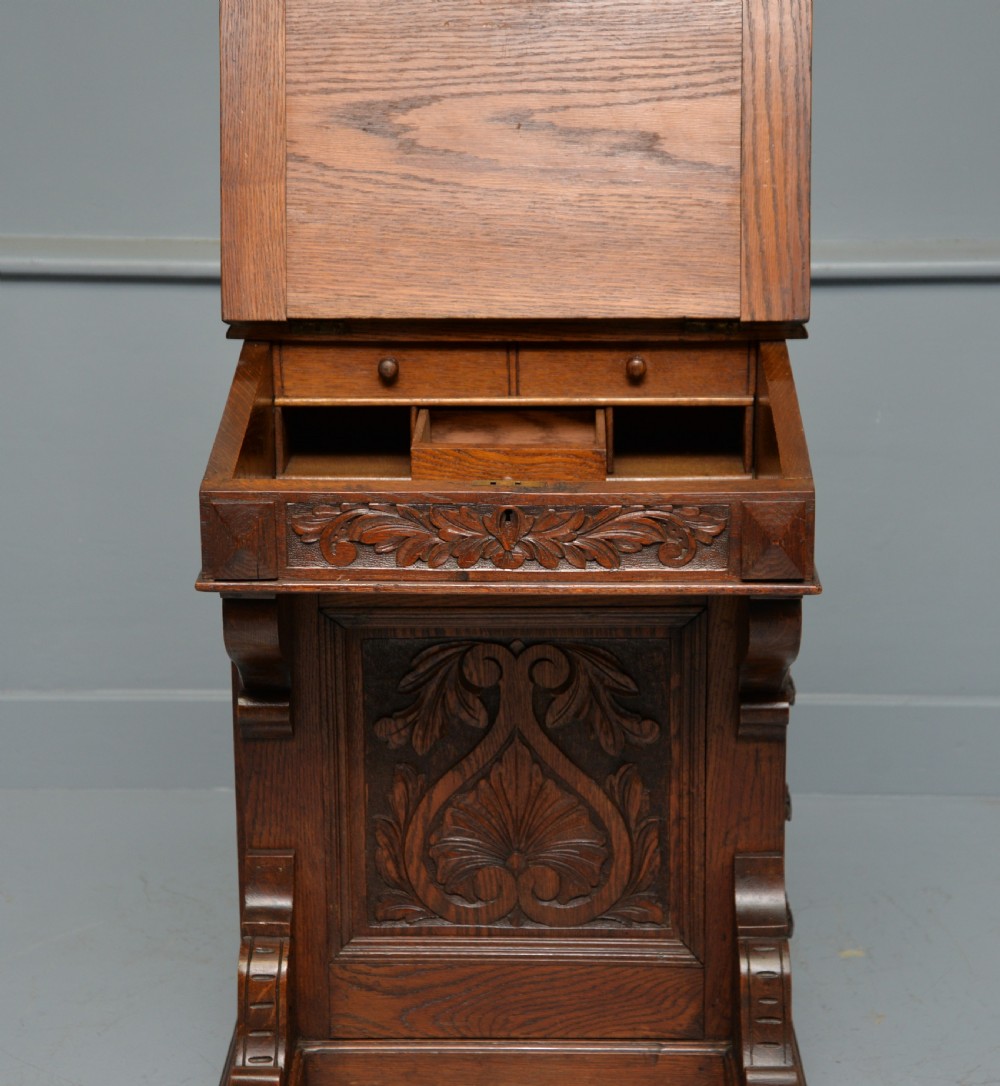 A Victorian dark carved oak Davenport desk featuring a red leather writing slant, carved Green Man mask handles on four side drawers, and a raised stationery gallery. View VII