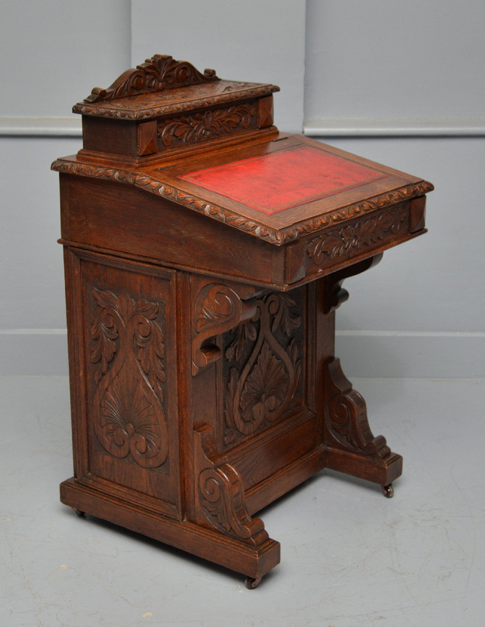 A Victorian dark carved oak Davenport desk featuring a red leather writing slant, carved Green Man mask handles on four side drawers, and a raised stationery gallery. View IX