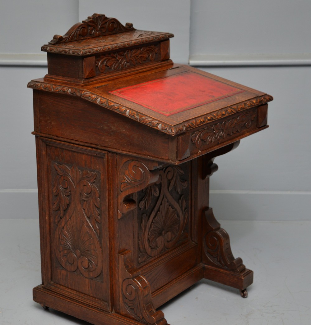A Victorian dark carved oak Davenport desk featuring a red leather writing slant, carved Green Man mask handles on four side drawers, and a raised stationery gallery. View X