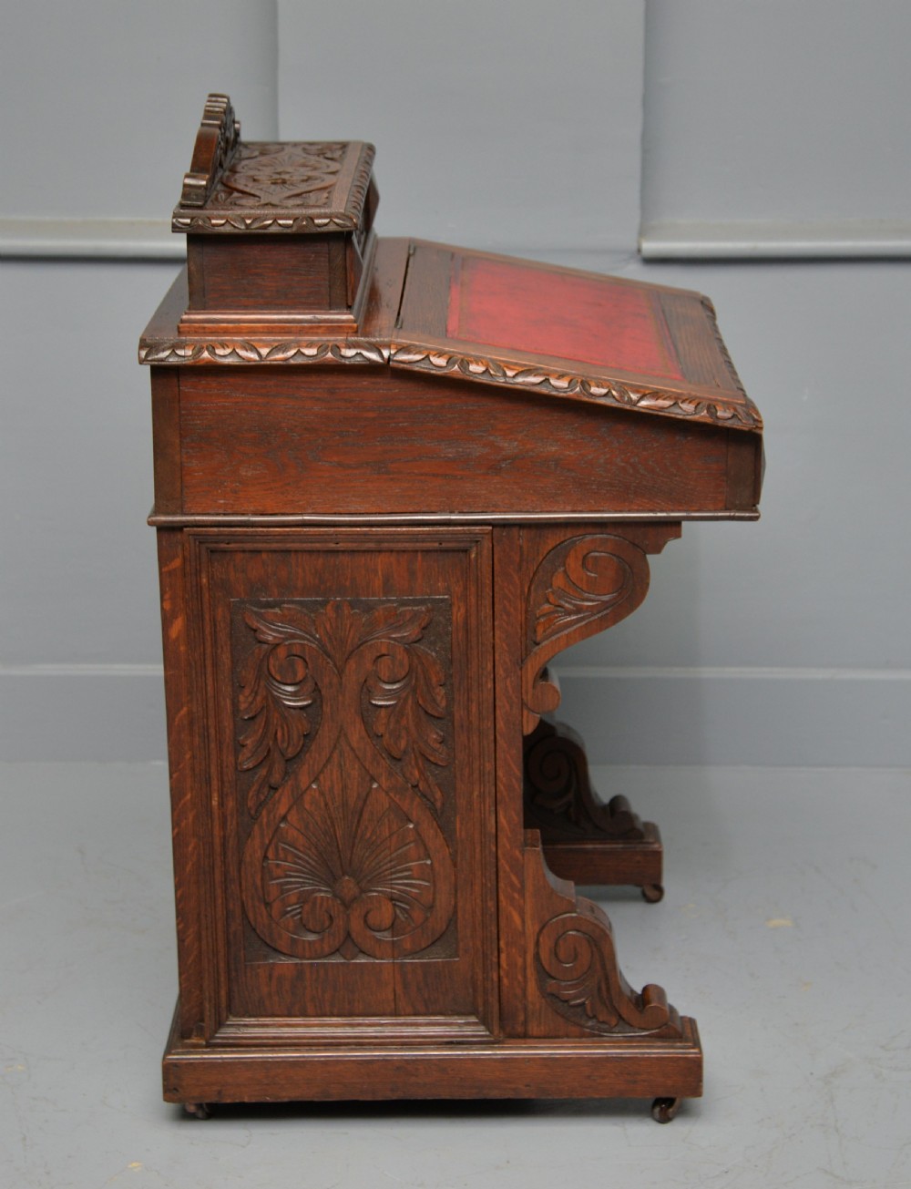 A Victorian dark carved oak Davenport desk featuring a red leather writing slant, carved Green Man mask handles on four side drawers, and a raised stationery gallery. View XI
