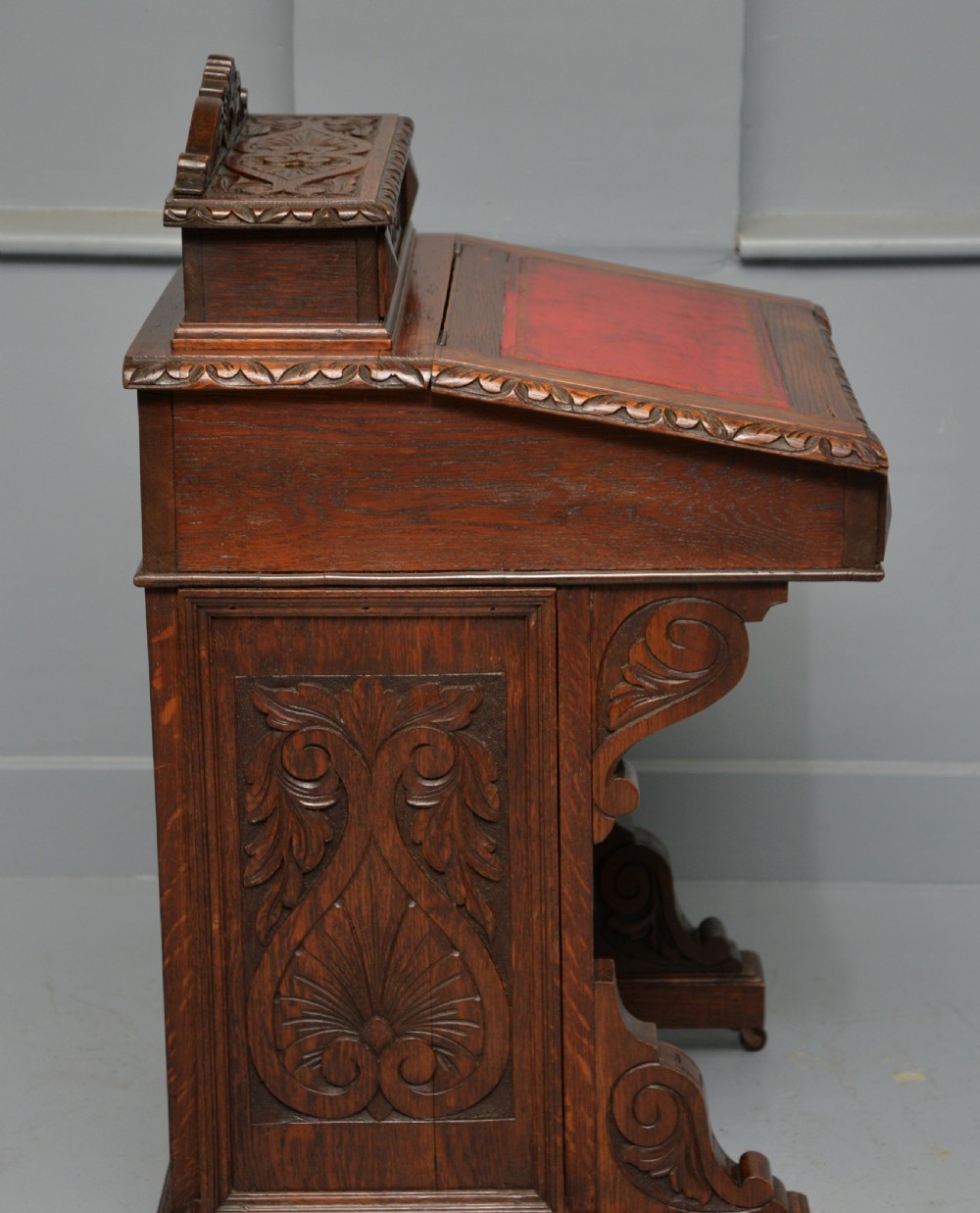A Victorian dark carved oak Davenport desk featuring a red leather writing slant, carved Green Man mask handles on four side drawers, and a raised stationery gallery. View XII