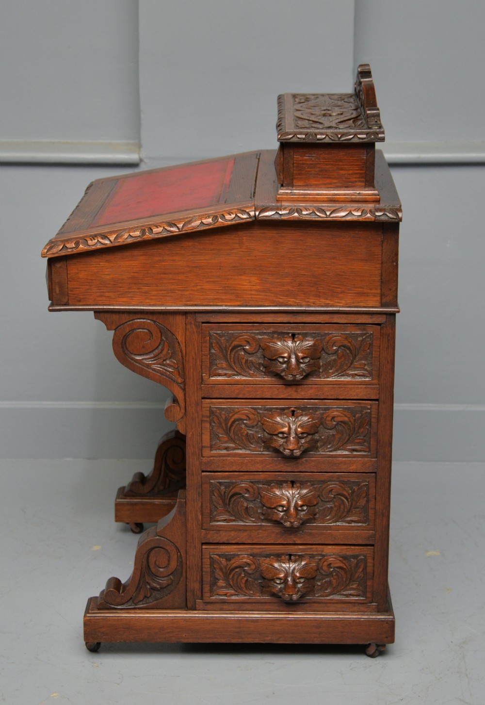 A Victorian dark carved oak Davenport desk featuring a red leather writing slant, carved Green Man mask handles on four side drawers, and a raised stationery gallery. View XV